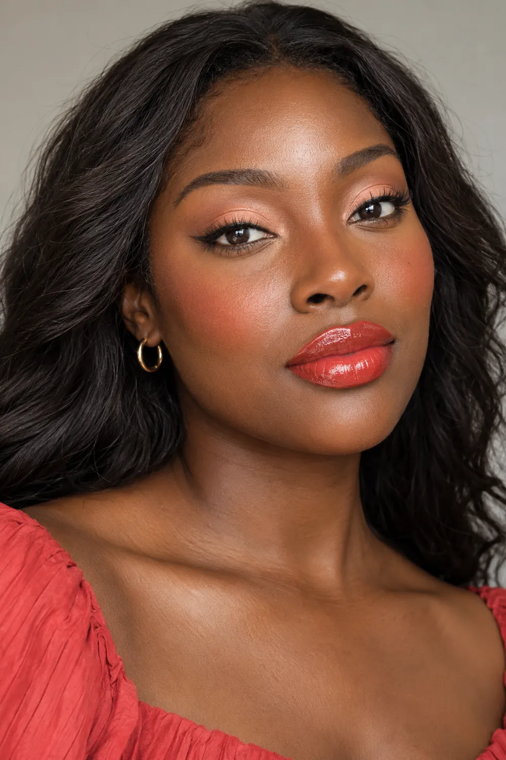 Adult woman with deep brown skin, glossy coral-red lips, peach eyeshadow, softly lifted brown liner, and high coral blush.