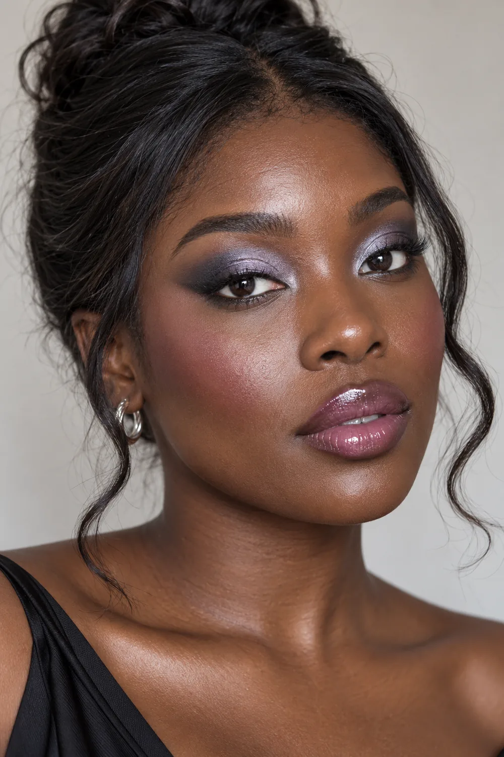 Adult woman with deep ebony skin wearing silver-lilac frosted eyeshadow, a diffused charcoal wing, mauve blush, and glossy plum lips.