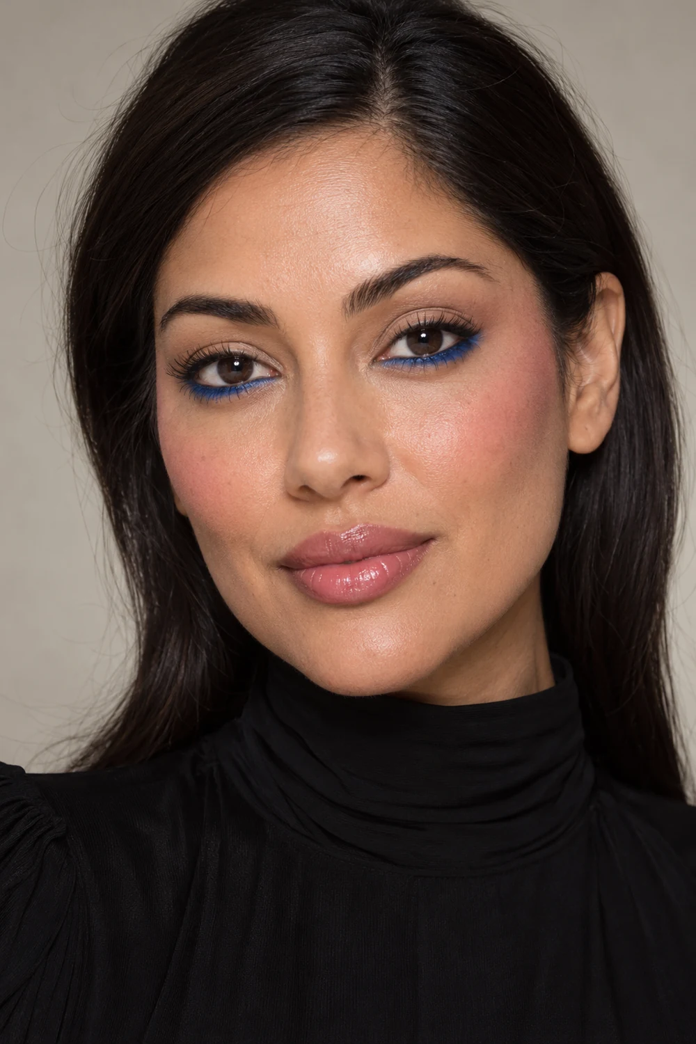 Close beauty portrait of an adult woman in a black high-neck dress, wearing cobalt lower-lash liner, soft sand eyeshadow, temple-placed pink blush, and glossy sheer pink lipstick.