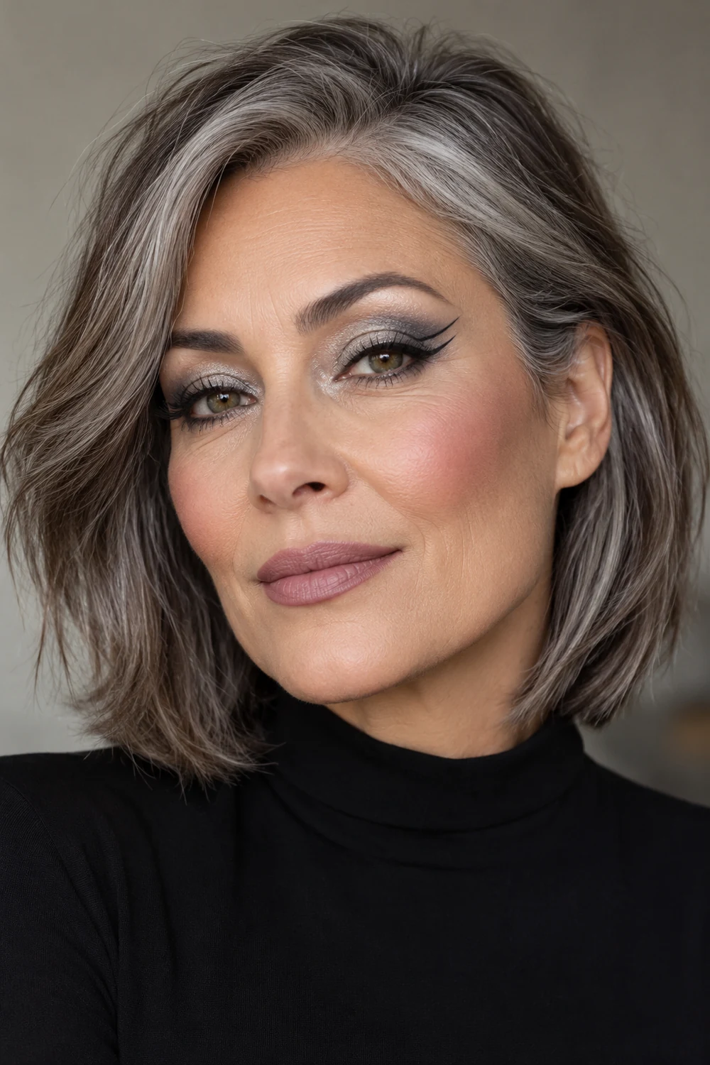 Mature woman with a salt-and-pepper bob wearing a high-neck black dress, silver-to-slate eyeshadow, separated double black wing liner, rose blush, and matte mauve lipstick.