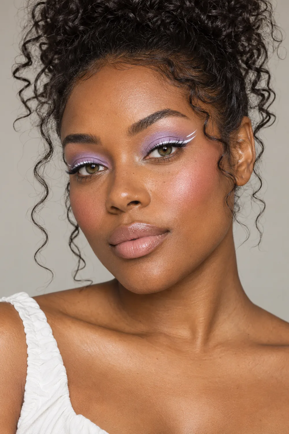 Beauty portrait of an adult Black woman with green-brown hazel eyes, lilac eyeshadow, crisp white double-point liner, cool pink blush, and pale satin nude lips.