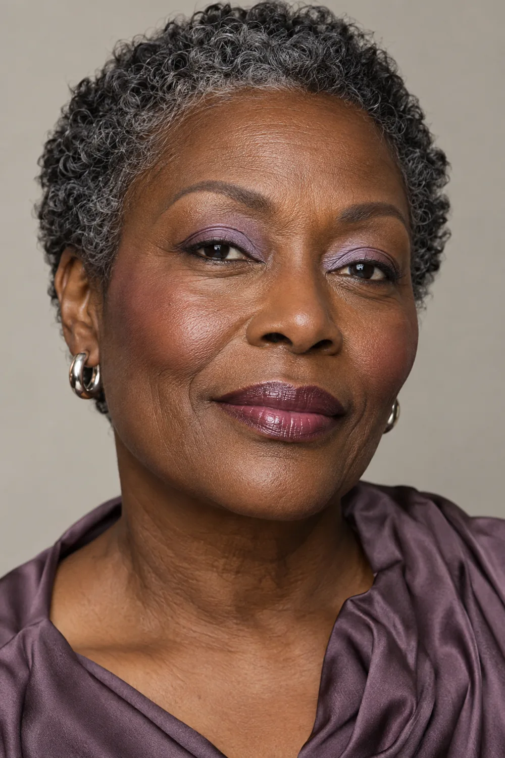 Mature woman with very deep brown skin wearing muted lavender inner-lid shadow, neutral rose blush high on her cheekbones, and glossy cool plum balm.