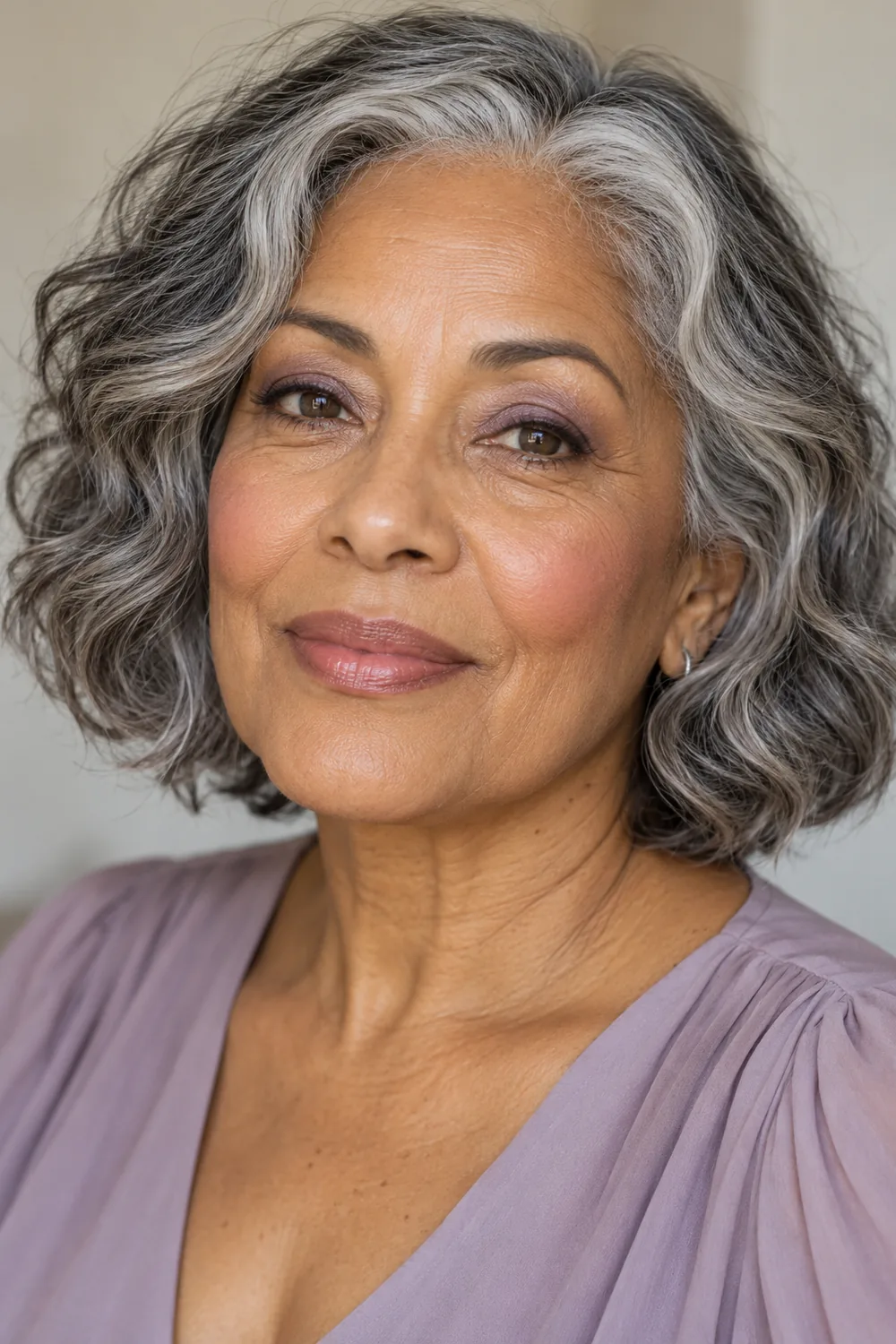 Fictional mature woman with light brown skin, a wavy silver-and-black bob, lavender and lilac eyeshadow, fine gray-brown liner, cool rose blush, and rosy nude satin lipstick.