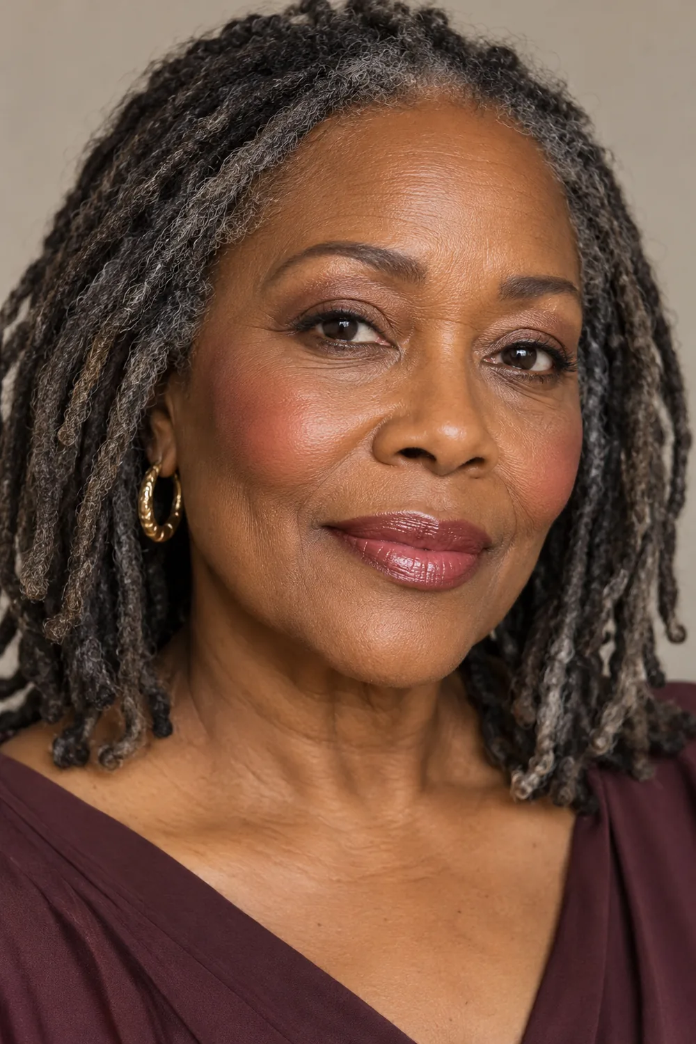 Portrait of a mature woman with deep medium skin and silver-threaded locs, wearing cream lid shadow, bronze lower-lash shading, soft berry blush, and glossy cherry-rose balm.
