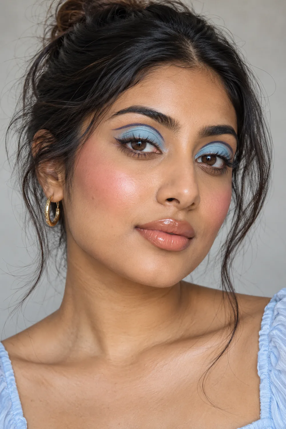 Adult South Asian woman wearing pastel baby-blue lid shadow with navy floating crease lines, pink blush, and peach nude gloss.