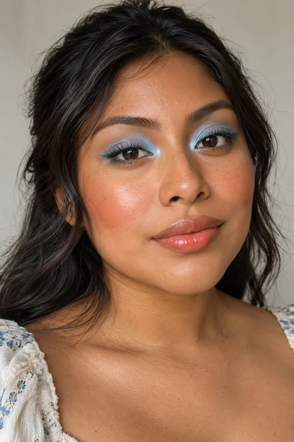 Adult woman with baby-blue eyeshadow, icy-blue inner corners, thin teal liner, cinnamon blush, and glossy coral lips.