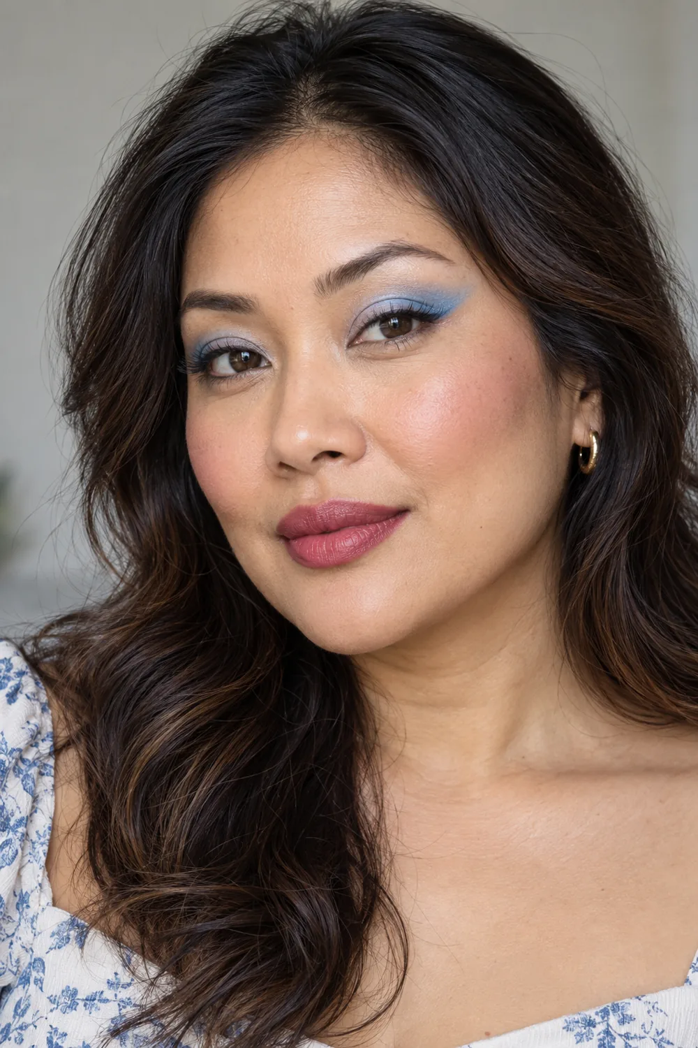 Close portrait of a woman wearing tapered pastel-blue eyeshadow wings, thin dark lash liner, pink blush, and a berry lip stain.