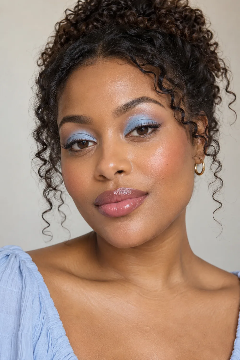 Adult Black woman wearing rounded powder-blue eyeshadow, fine brown liner, apricot blush, and glossy rose lipstick.
