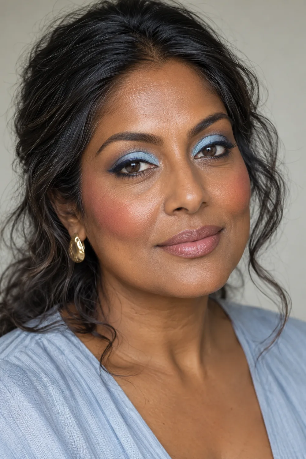Adult South Asian woman with baby-blue lid shadow, softly smudged navy upper lash liner, berry-rose blush, and satin beige-pink lipstick.