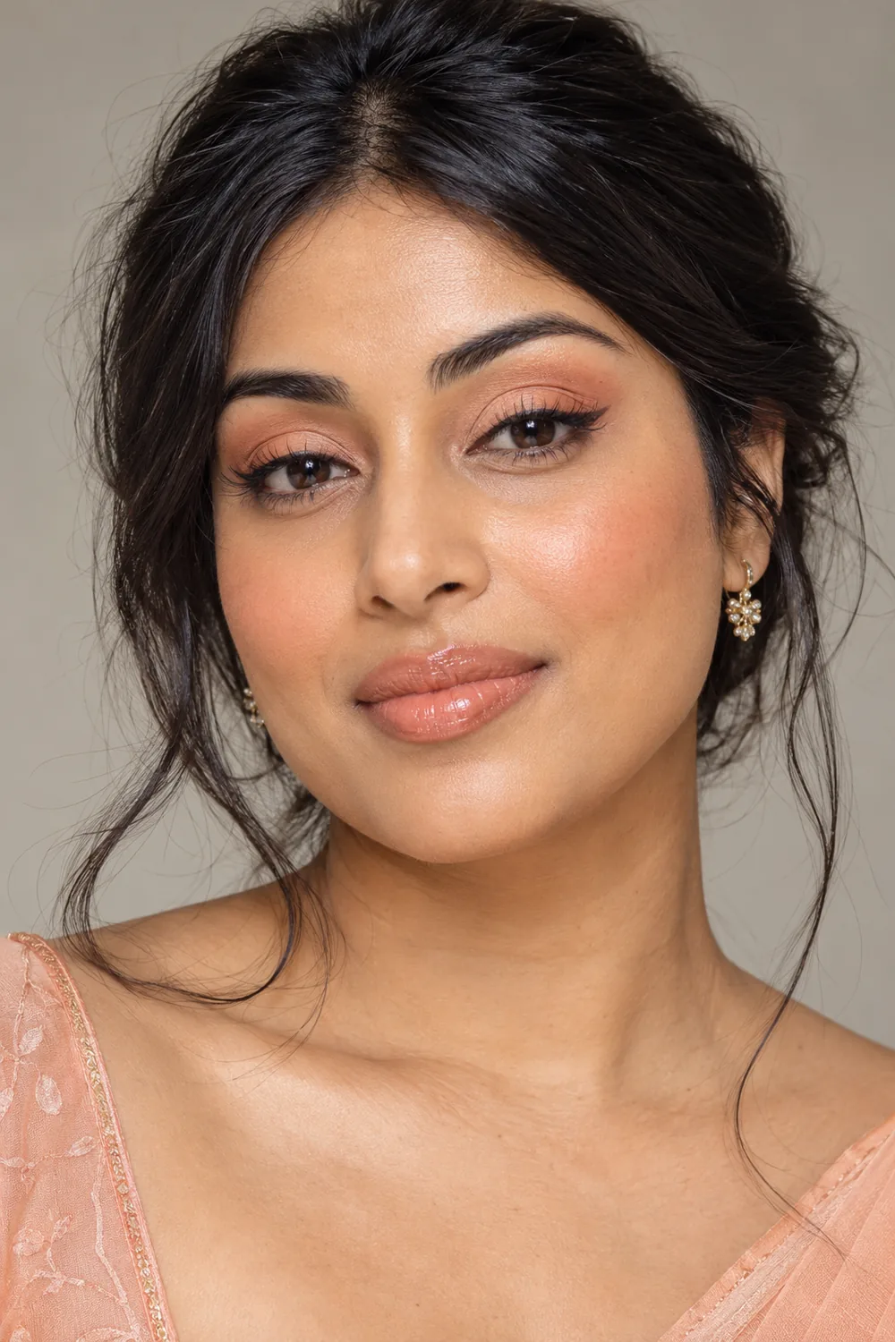 Adult South Asian woman with warm peach and apricot eye makeup, softly winged brown liner, rounded peach blush, and glossy nude-peach lips.