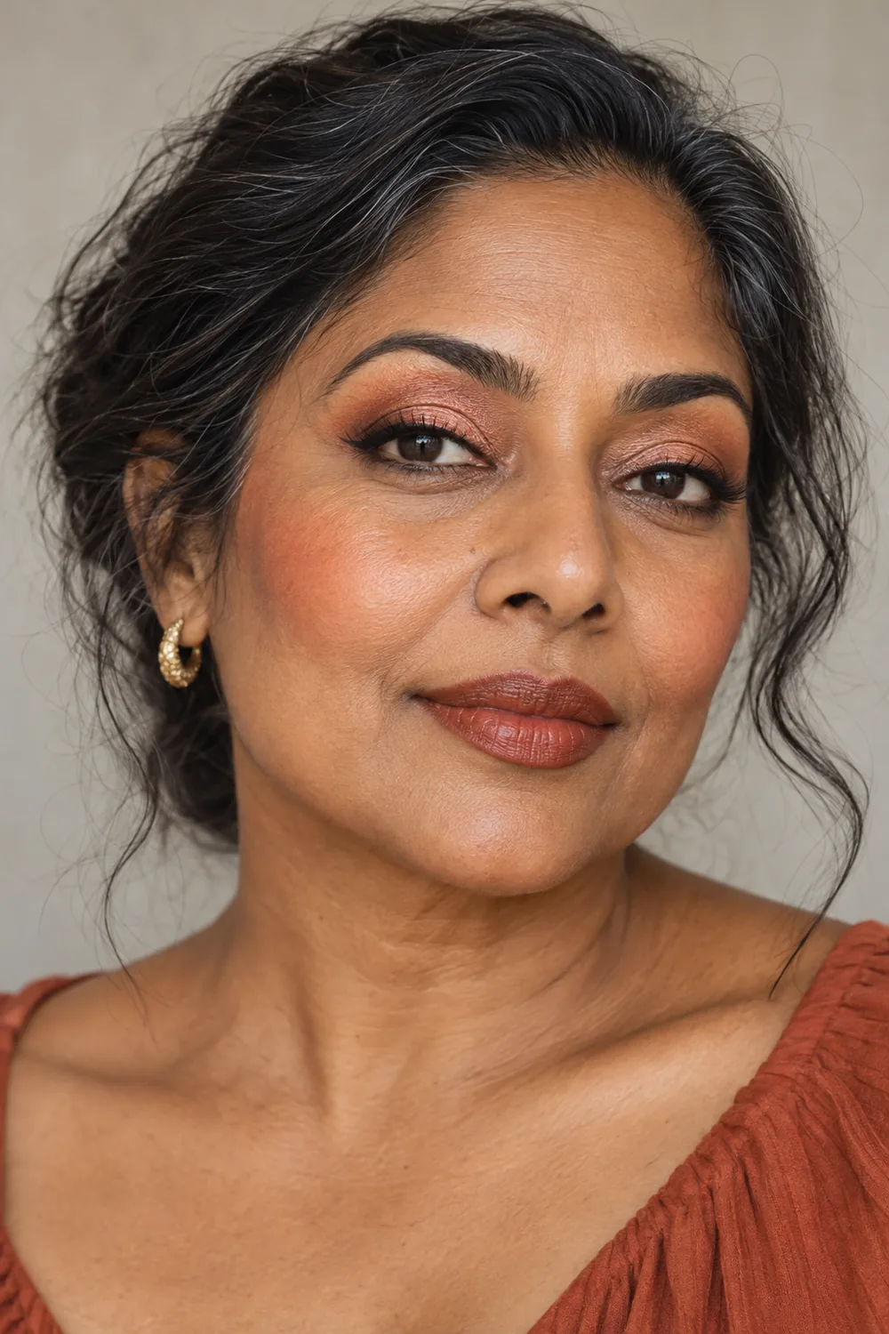 Adult South Asian woman with copper-peach metallic eyeshadow, a warm brown wing, peach cheekbone blush, and deep cinnamon satin lipstick.