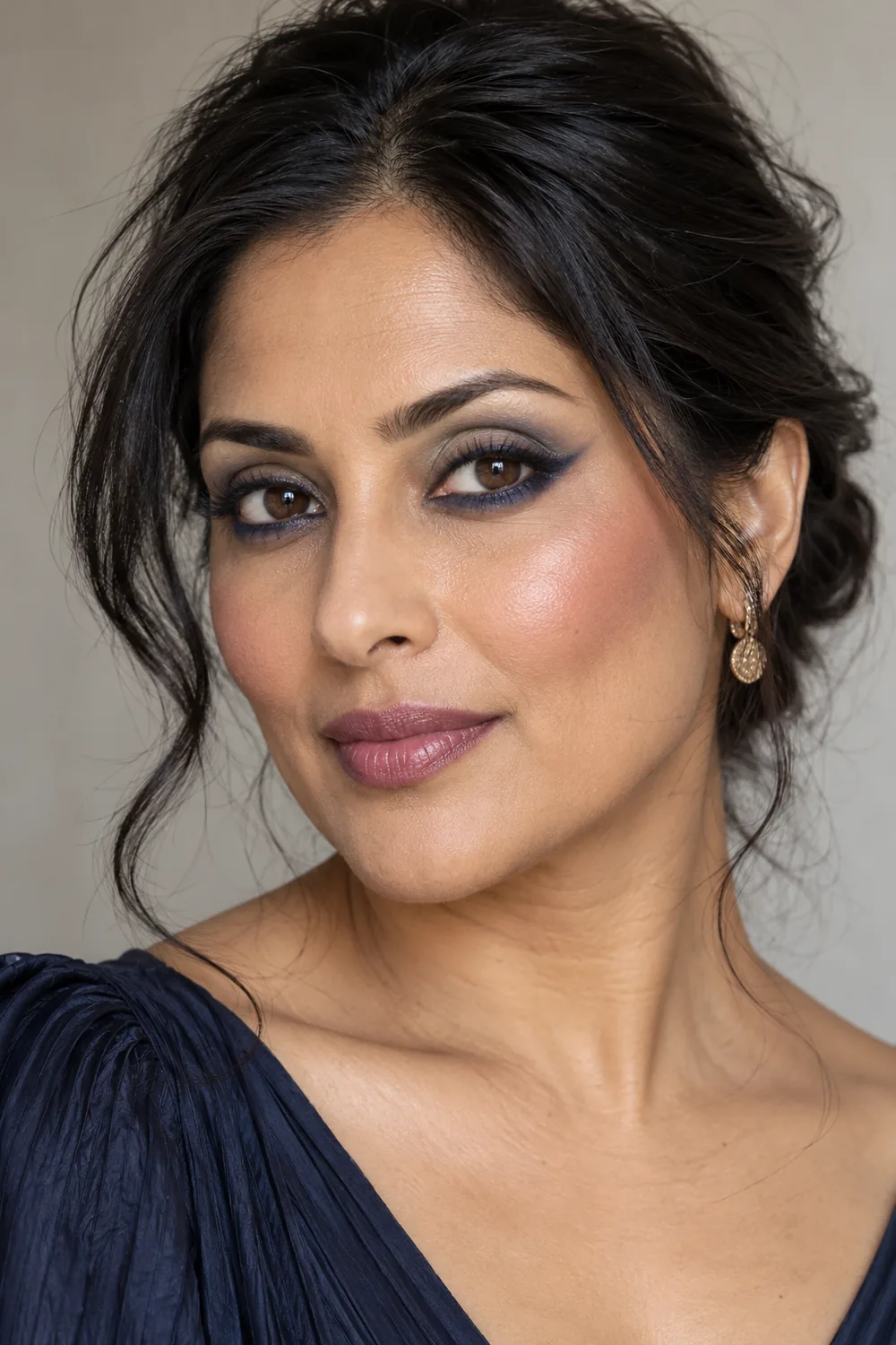 Adult South Asian woman with open dark eyes, navy lower-lash liner extending into tapered outward wings, muted gray-blue shadow, plum blush, and mauve satin lipstick.