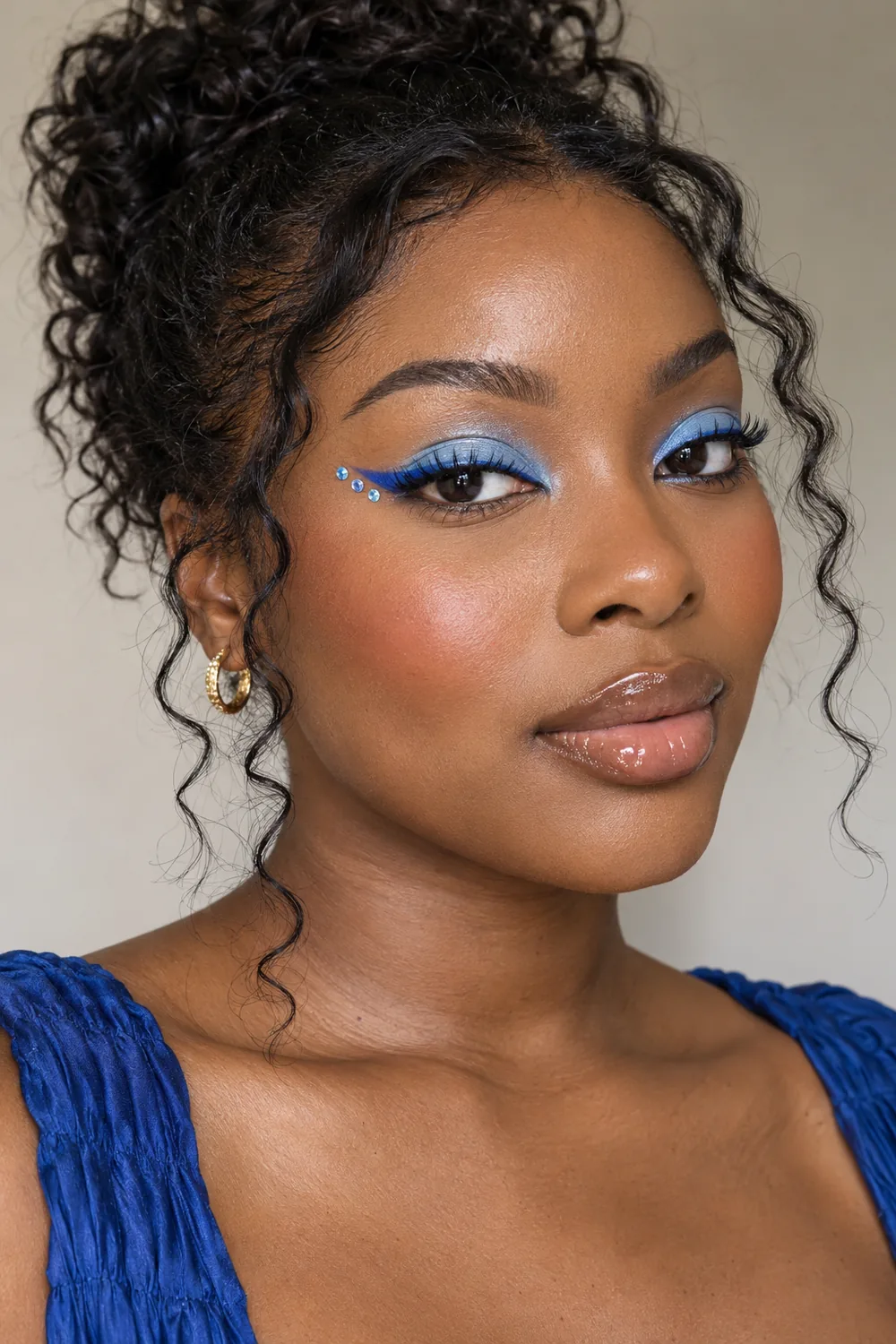 Adult woman with deep brown skin wearing icy blue lid color, a cobalt graphic wing, three blue-clear gems outside the liner tip, peach blush, and glossy nude lips.