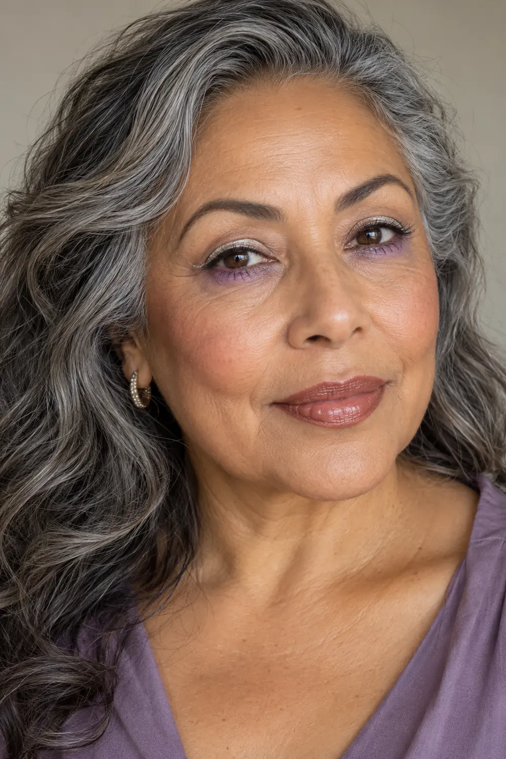 Close portrait of an adult woman with salt-and-pepper hair, lavender lower-lid shadow, narrow silver upper liner, warm nude blush, and creamy rosewood lips.