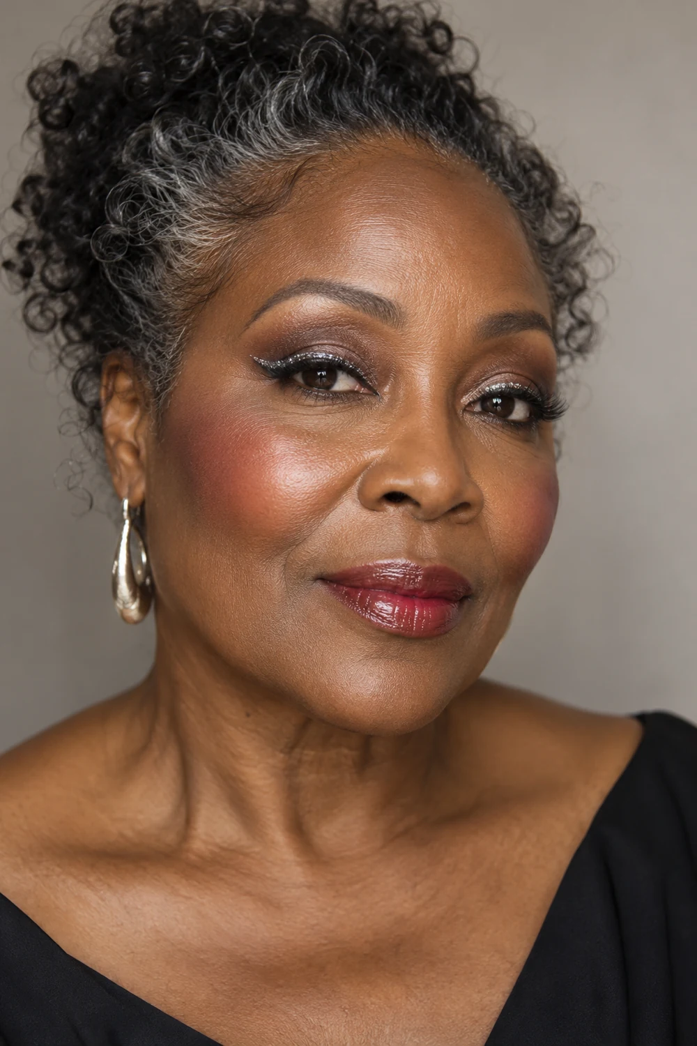 Close portrait of an adult Black woman with silver strands in her curls, smoky brown eye shadow, hooked silver eyeliner, pink-red blush, and glossy cherry lips.