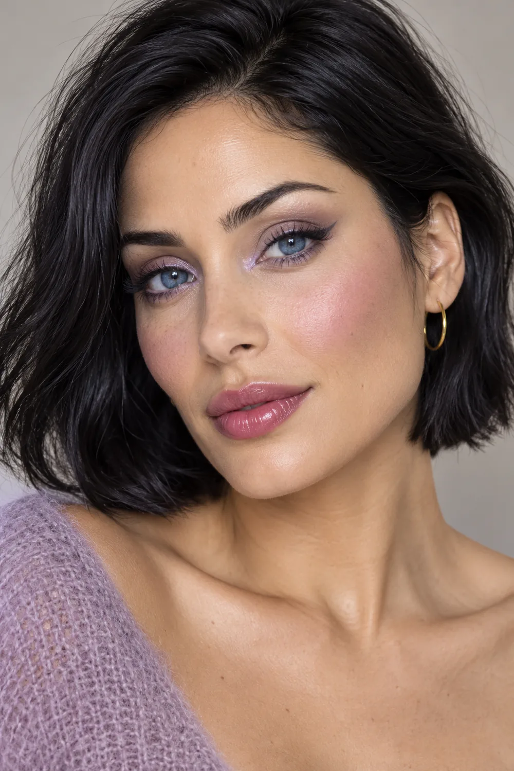 Adult woman with blue eyes and a black bob wearing taupe eyeshadow, lilac inner-corner shimmer, charcoal winged liner, lavender-pink blush, and berry-tinted glossy lips.