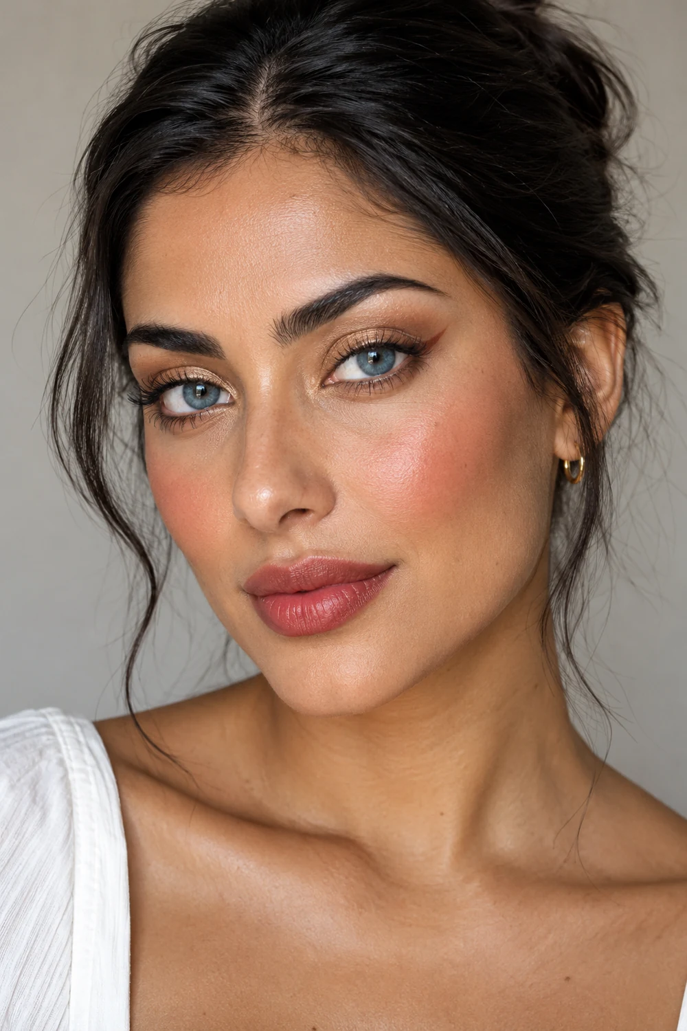Close portrait of an adult woman with blue eyes, dark hair pulled back, soft gold shimmer shadow, a small brown outer-corner flick, peach blush, and muted red satin lipstick.